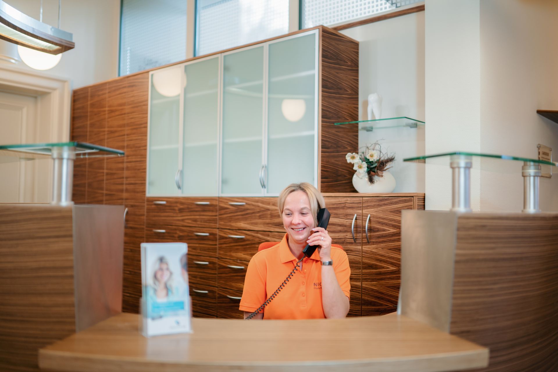 Frau sitzt lächelnd an der Anmeldung hinter dem Empfangstresen und telefoniert